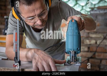 Un charpentier caucasien se concentre sur la coupe du bois avec une scie sauteuse dans son atelier. Banque D'Images