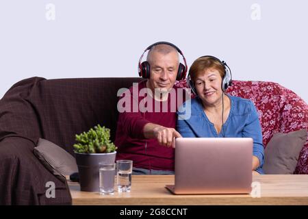 Les personnes âgées assises sur un canapé, toutes deux portant un casque, parlent en ligne avec quelqu'un sur l'ordinateur. Banque D'Images
