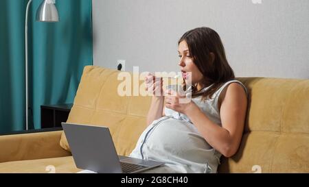 Une jeune femme enceinte mange du yaourt et regarde un ordinateur portable tout en étant assise sur un canapé. Banque D'Images