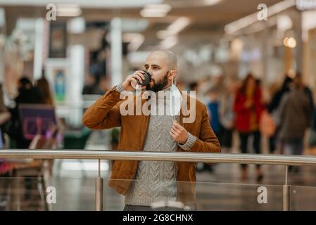 Un homme boit du café tout en prenant un masque médical dans le centre commercial. Un homme chauve garde la distance sociale. Banque D'Images