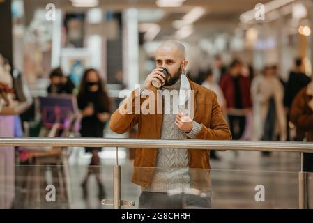 Un homme tient un masque médical à l'entaille tout en buvant du café dans le centre commercial. Un homme chauve garde la distance sociale. Banque D'Images