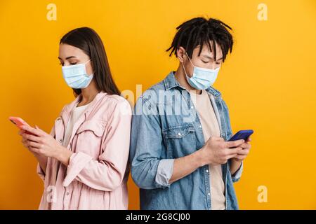 Jeune couple multiracial dans des masques de visage utilisant des téléphones mobiles isolés sur le mur jaune Banque D'Images