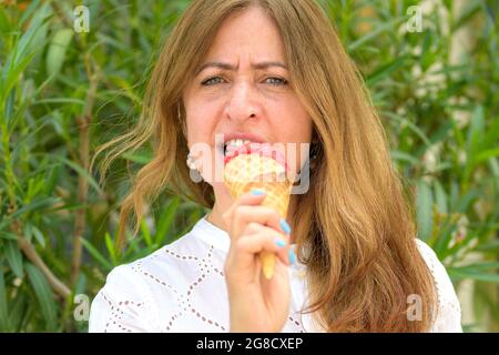 Femme frognant à l'appareil photo comme elle a le goût de la glace de baie dans un cône de sucre à l'extérieur contre la verdure en été dans un portrait étroit Banque D'Images