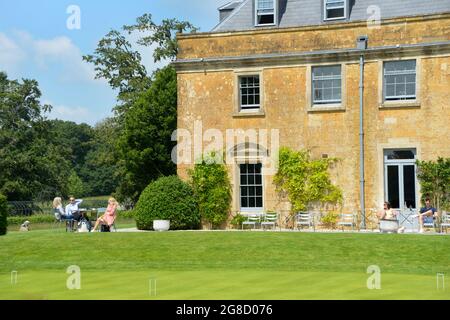 Les gens glamour s'assoient sur le patio en face d'une belle maison géorgienne appelée Maison Hadspen dans le Somerset. Il fait partie du nouvel hôtel Newt près de Bruton. Une pelouse de croquet repose au premier plan. L'image reflète une belle maison de campagne anglaise classique avec des gens qui l'apprécient au soleil. Banque D'Images