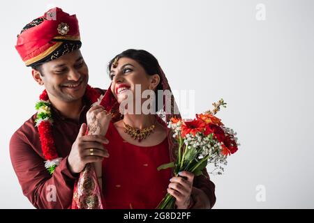 homme indien avec anneau sur le doigt tenant la main de la mariée heureuse avec bouquet de fleurs isolées sur blanc Banque D'Images