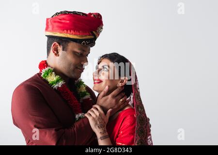 un indien heureux touchant le visage d'une mariée gaie en foulard et sari isolé sur gris Banque D'Images