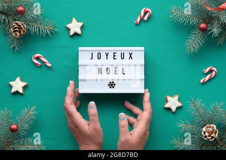 Boîte lumineuse avec texte Joyeux Noël - Joyeux Noël en langue française. Les mains tiennent la boîte. Arrière-plan de Noël, vue de dessus sur les brindilles de sapin avec des décorations rouges Banque D'Images