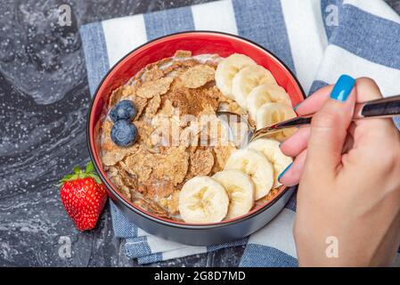 Muesli. Petit déjeuner, alimentation saine et alimentation. Muesli au lait et aux fruits dans une assiette sur un dessus en marbre noir. Main de femme avec une cuillère Banque D'Images