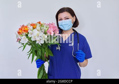 Femelle medic en bleu uniforme masque médical avec bouquet de fleurs sur fond clair Banque D'Images