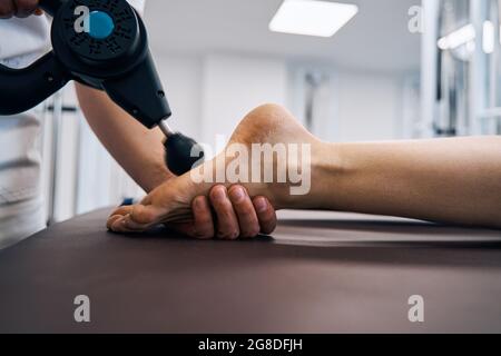 Gros plan du pied de femme de massage frottant avec dispositif de percussion de massage dans la salle de thérapie des cliniques de réadaptation Banque D'Images