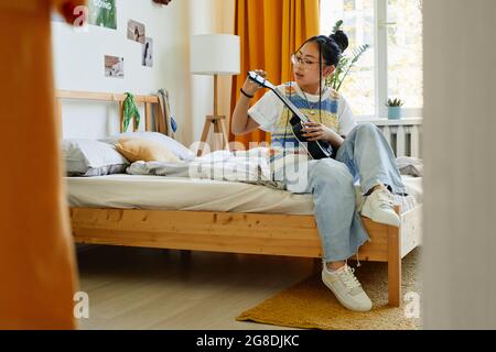 Portrait complet d'une adolescente tendance jouant ukulele tout en étant assise sur le lit dans une chambre confortable Banque D'Images