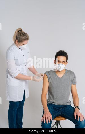 Cliché studio d'un jeune homme portant un masque facial recevant un vaccin contre le coronavirus assis sur une chaise sur un fond blanc isolé. Banque D'Images