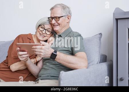 Portrait minimal d'un couple âgé moderne utilisant une tablette à la maison et souriant avec joie en étant assis sur un canapé, dans un espace de copie Banque D'Images