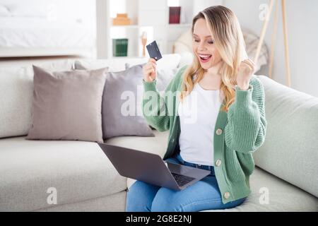 Portrait d'une jolie fille à cheveux ondulés et tendance assise sur un divan en utilisant un ordinateur portable pour commander des achats maison lumière blanc plat à l'intérieur Banque D'Images