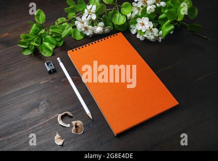 Photo d'un modèle de papeterie vierge. Carnet à croquis, crayon, aiguiseur et fleurs printanières orange sur fond de table en bois. Banque D'Images