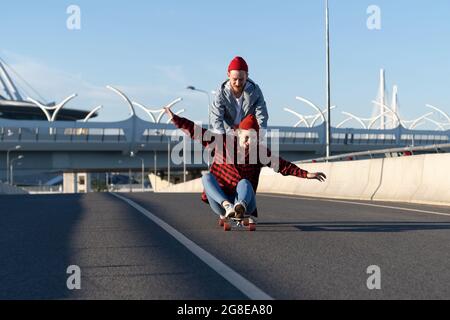 Décontracté couple sur le long-board: Homme enseignant fille à l'équilibre sur le skateboard la pousser en arrière sur la route vide Banque D'Images