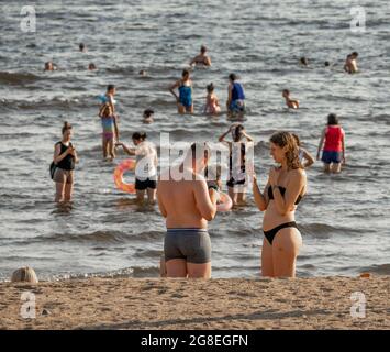 Les jeunes hommes et femmes en maillot de bain vérifient leurs téléphones sur la plage par le golfe finlandais avec des personnes de baignade, Saint-Pétersbourg, Russie Banque D'Images