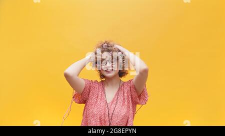 Belle fille portant des lunettes de nerdy avec les deux mains sur sa tête et souriant. Fille d'une humeur joyeuse et portant des écouteurs à l'écoute de sa musique préférée. Isolé sur fond jaune vif. Banque D'Images