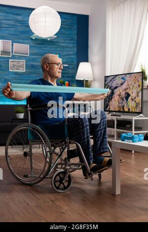 Homme âgé handicapé en fauteuil roulant avec bande élastique exercice d'entraînement de corps se rétablissant après un accident d'invalidité regardant la vidéo de stretch sur tablette. Retraité faisant de l'exercice de bras de soins de santé Banque D'Images