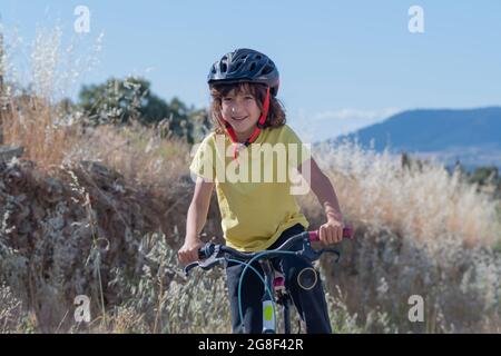 jeune garçon souriant avec de longs cheveux et un casque de sécurité a l'amusement de conduire son vélo à travers les rues de son village en été lors d'une journée ensoleillée Banque D'Images