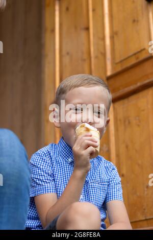 Un enfant gai content dans une chemise bleue mange de la glace sur le porche d'une maison dans un village par une journée ensoleillée d'été. Mise au point sélective. Portrait Banque D'Images