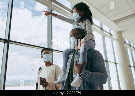 Père avec fille sur l'épaule et pointant des avions tout en marchant avec la mère à l'aéroport. La famille africaine à l'aéroport en pandémie. Banque D'Images
