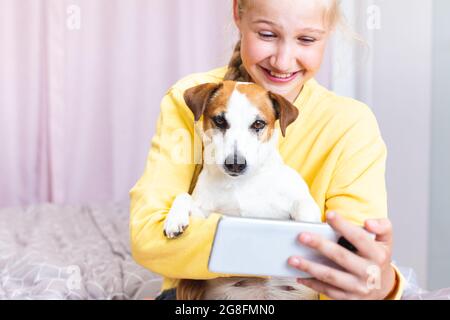 Une fille qui rit avec un téléphone portable dans sa main, avec son chien Jack Russell Terrier, communique sur les médias sociaux ou via Zoom, fait un selfie Banque D'Images