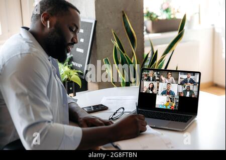 E-learning. Un gars afro-américain concentré est assis à la table à la maison, écoute une leçon en ligne par ordinateur portable, prend des notes, sur un écran une femme enseignant montre des informations sur un tableau blanc, et d'autres étudiants Banque D'Images