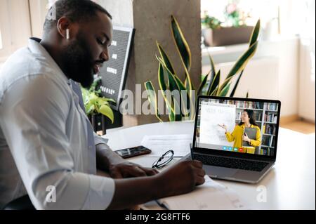 Formation en ligne. Un homme afro-américain concentré est assis à la table à la maison, écoute une leçon en ligne par ordinateur portable, prend des notes, sur l'écran une femme enseignant montre des informations sur un tableau blanc, appel vidéo Banque D'Images