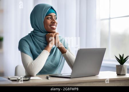 Femme noire musulmane pensive heureuse dans hijab assis au bureau avec ordinateur portable Banque D'Images