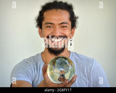 Beau jeune hispanique Latino-homme avec anneau de nez tient une boule de cristal transparente avec son image miroir dans sa main droite et sourit au spectateur. Banque D'Images