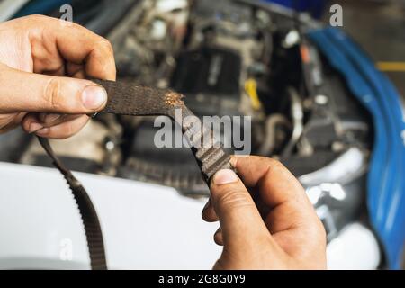 Un mécanicien d'automobiles montre une courroie de distribution déchirée avec des dents usées sur le fond d'un gros plan de capot de voiture ouvert Banque D'Images