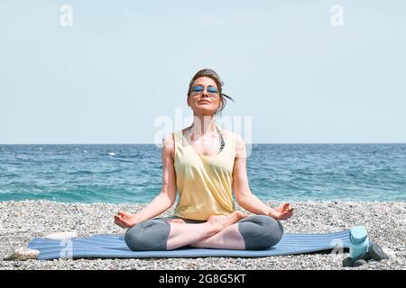 Femme, assise à Padmasana ou lotus pose, pratique le yoga sur la plage le matin, elle médite, regardant sur la mer bleue. Concept de bien-être. Banque D'Images