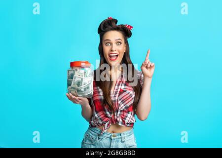 Concept d'économie d'argent. Jeune femme joyeuse pinup tenant un pot de dollars, pointant vers le haut sur fond bleu de studio Banque D'Images