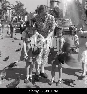 Années 1950, historique, mère avec ses deux jeunes filles, nourrissant les pigeons de Trafalgar Square, Londres, Angleterre, Royaume-Uni. Une activité traditionnelle depuis plus de cent ans, des troupeaux de pigeons descendraient sur la place pour recevoir la nourriture de ceux qui visitaient le célèbre centre de Londres. Banque D'Images