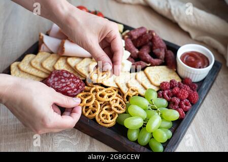 Cuisine de fête, les mains des femmes posant le mini bretzel sur la carte de charcuterie avec saucisse, fruits, crackers et fromage, gros plan. Banque D'Images