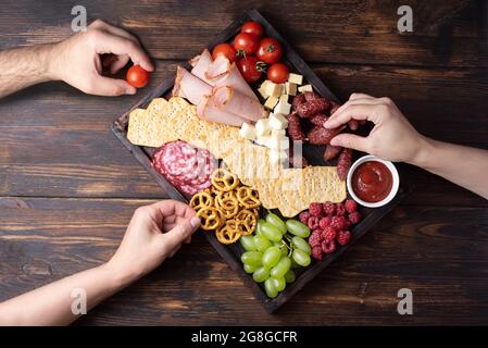 Mains de femmes et de hommes prenant des en-cas à la carte de charcuterie avec saucisses, fruits, crackers et fromage sur un fond en bois sombre, gros plan. Banque D'Images