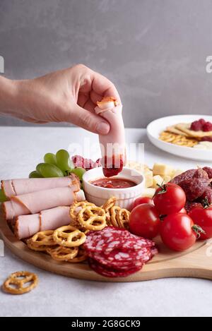 Assiettes avec hors-d'œuvre, une femelle trempant une tranche de jambon dans une sauce sur un plateau de charcuterie rond avec saucisse, fromage, crackers et fruits, gros plan. Banque D'Images