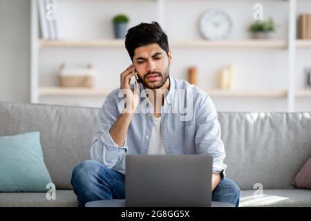 Activités à distance. Inquiet indépendant arabe parlant sur téléphone portable et utilisant ordinateur portable, assis sur un canapé à la maison Banque D'Images