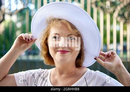 Belle femme âgée de 54 ans en chapeau blanc sourit tout en marchant dehors. Banque D'Images