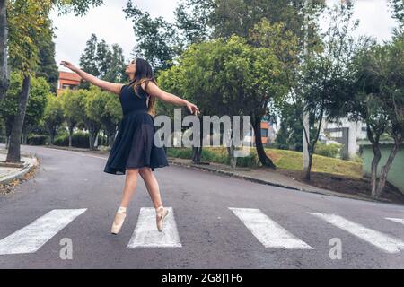 Jeune danseuse latine avec robe noire marchant avec élégance avec des chaussures de ballet sur une traversée piétonne dans la rue Banque D'Images