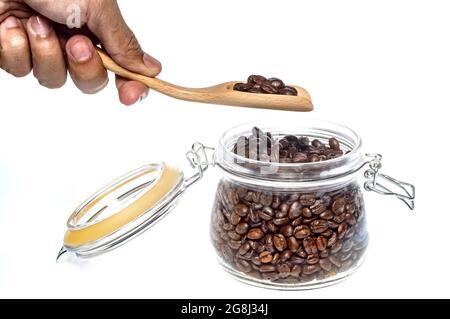 main d'un homme tenant une cuillère en bois pour ramasser des grains de café torréfiés dans un pot en verre sur fond blanc Banque D'Images