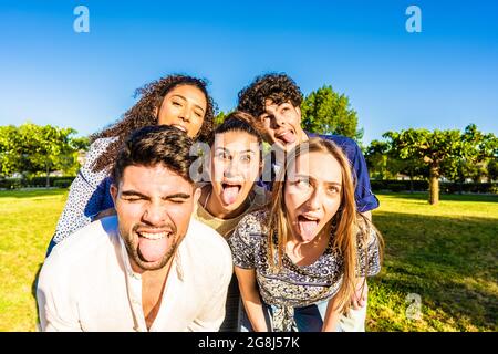 Groupe de jeunes idiot multiracial millénaires amis faisant des visages drôles avec la langue, la bouche ouverte, et les yeux qui posent pour un portrait dans le parc de la ville. Banque D'Images