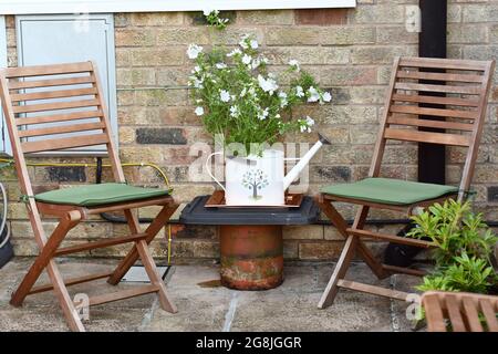 Une table de fortune d'un pot de cheminée tenant un arrosoir avec des géraniums sauvages et deux chaises vides Banque D'Images