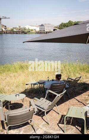 Une femme bénéficie de la vue assise à une table sur les rives de la Loire avec une bouteille d'eau, à Trentemoult, en France Banque D'Images