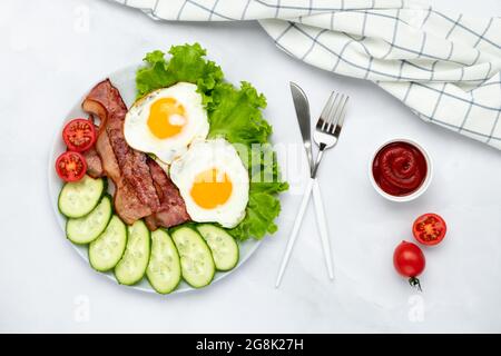 Œufs frits avec bacon et légumes sur une table grise, nourriture de base le matin. Concombres et tomates frais. Concept petit déjeuner. Vue de dessus. Pose à plat Banque D'Images