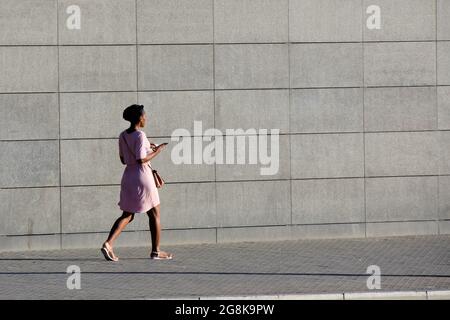 La jeune femme noire marche près d'un mur gris dans des vêtements d'été pendant qu'elle tient son smartphone. Banque D'Images