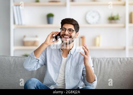 Joyeux homme arabe émotionnel ayant la conversation téléphonique à la maison, a obtenu de bonnes nouvelles, assis sur le canapé dans le salon Banque D'Images