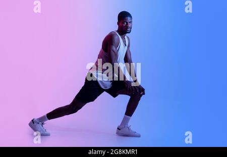 Pleine longueur d'homme noir sportif faisant des fentes ou étirant ses jambes en lumière de néon. Concept de mode de vie actif Banque D'Images