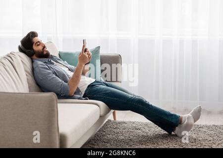 Détente à l'intérieur. Jeune garçon arabe avec casque sans fil et smartphone reposant sur un canapé confortable à la maison, vue latérale Banque D'Images